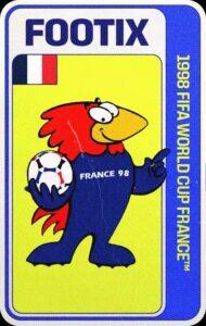 Footix, mascota oficial del Mundial Francia 1998, que representa a un gallo antropomórfico y caricaturesco de diseño minimalista, con cresta y cola de color rojo, pico amarillo, y un cuerpo y alas de color azul, así como las patas, que tienen forma de botines. Sobre el pecho, lleva la leyenda "France 98", y, mientras señala con un dedo hacia un lado, con la mano o ala opuesta, sujeta un balón de fútbol.