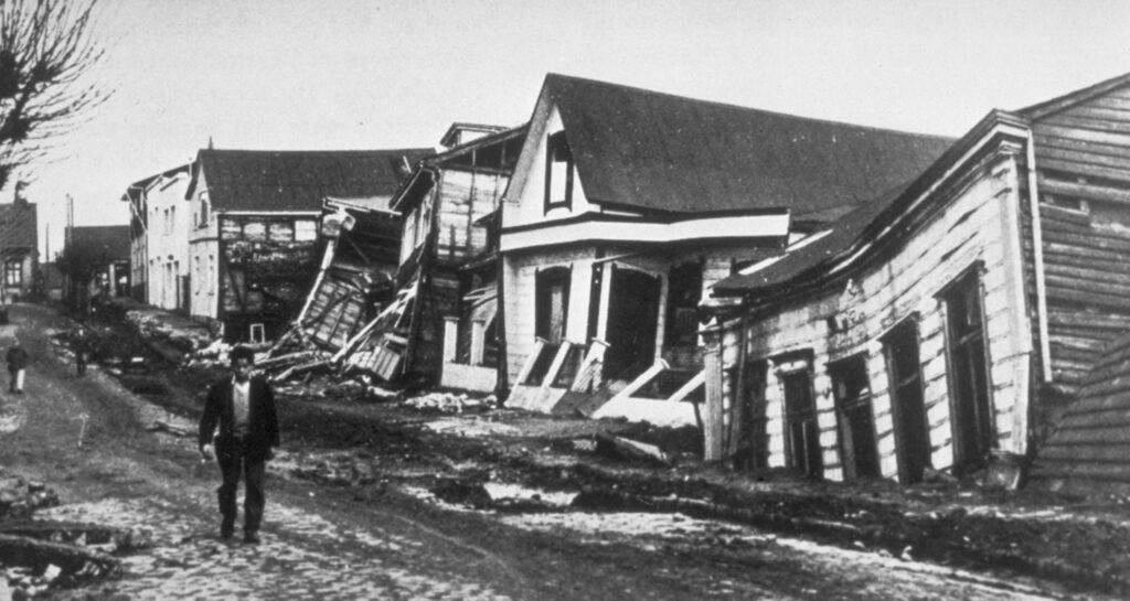 Fotografía en blanco y negro que muestra una calle en la ciudad de Valdivia, Chile, en el año 1960, en la que se muestra el daño producido a las casas de madera que se encuentran en ella tras el terremoto y tsunami de 9.5 en la escala de Richter, que tuvo lugar el 22 de mayo de 1960.