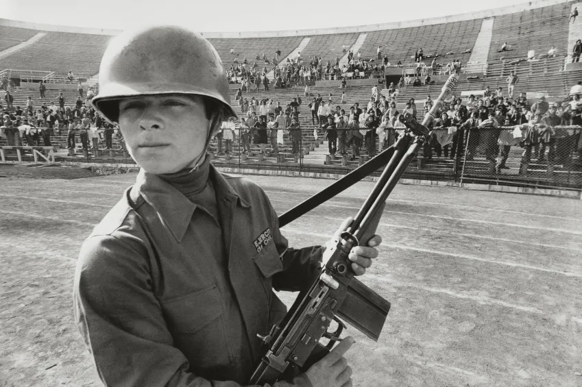 Un soldado posa con una metralleta en las pistas de atletismo del interior del Estadio Nacional de Chile; en el fondo de la imagen, se puede ver, repartidos por distintos puntos del graderío y separados por una reja baja de alambre del centro del estadio, a prisioneros llevados allí tras el golpe de estado llevado a cabo por Pinochet contra Salvador Ayende el 11 de septiembre de ese mismo año.