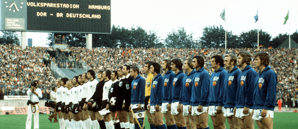 En una fila recta en mitad del campo, los jugadores de las selecciones de Alemania Federal (Alemania Occidental) y de la República Democrática de Alemania (Alemania Oriental) escuchan los himnos nacionales mientras flanquean a los árbitros del partido a punto de celebrarse en el Volksparkstadion de Hamburgo el 22 de junio de 1974.