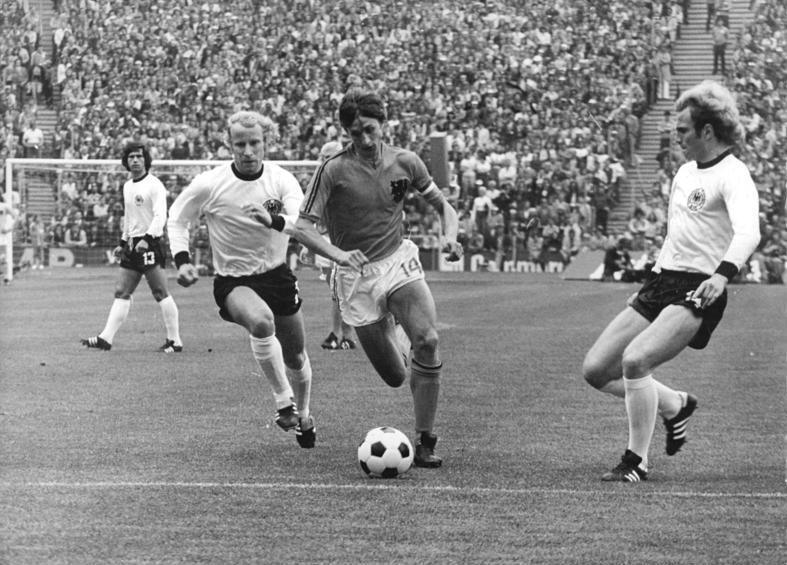 El jugador neerlandés Johannes Cruyff (14), controla el balón durante la final de la Copa del Mundo de la FIFA Alemania 1974 en el Olympiastadion de Múnich, mientras lo persiguen los jugadores de la selección de Alemania Federal Berti Vogts (2) a la izquierda y Uli Hoeness (14) a la derecha. Al fondo a la izquierda de la imagen, observando el desarrollo de la jugada, el jugador alemán Gerd Müller (13).