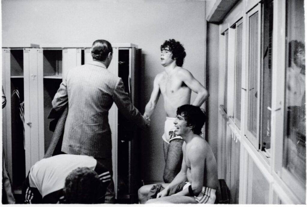 En el vestuario, después de haber ganado el partido contra Perú, Alberto Tarantini, en pantalones cortos y sin camiseta, da la mano recién sacada del calzón a Rafael Videla, que se encuentra de espaldas y vestido de traje. Sentado en el banco, en primer plano, el capitán Daniel Passarella, también sin camiseta y con cara de extenuación, mientras una persona cuya cara no se ve se agacha para recoger algo del suelo.