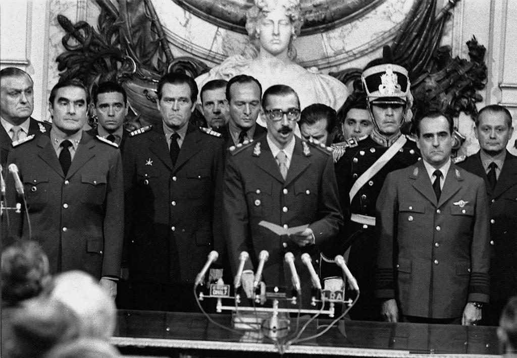 En el centro de la imagen, el general Rafael Videla, detrás de un escrtorio con seis micrófonos, con una hoja de papel en la mano, jura su cargo como presidente de la República Argentina en el Salón Blanco de la Casa Rosada (Buenos Aires), dando comienzo a la dictadura cívico-militar encabezada por este. Detrás de Videla, se encuentran los demás miembros de la Junta Militar.